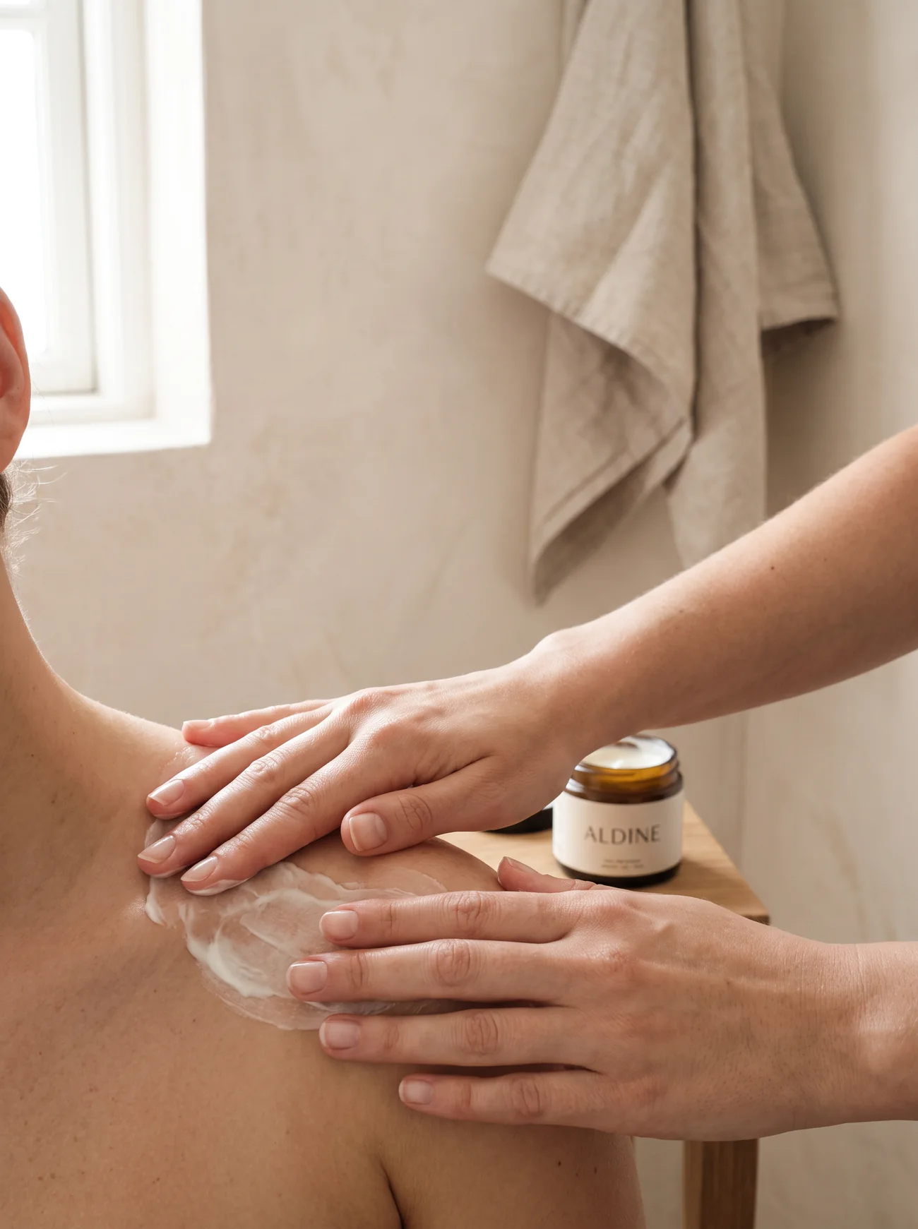 A therapist's bare hands smoothing cream over a client's shoulder in a calm treatment room