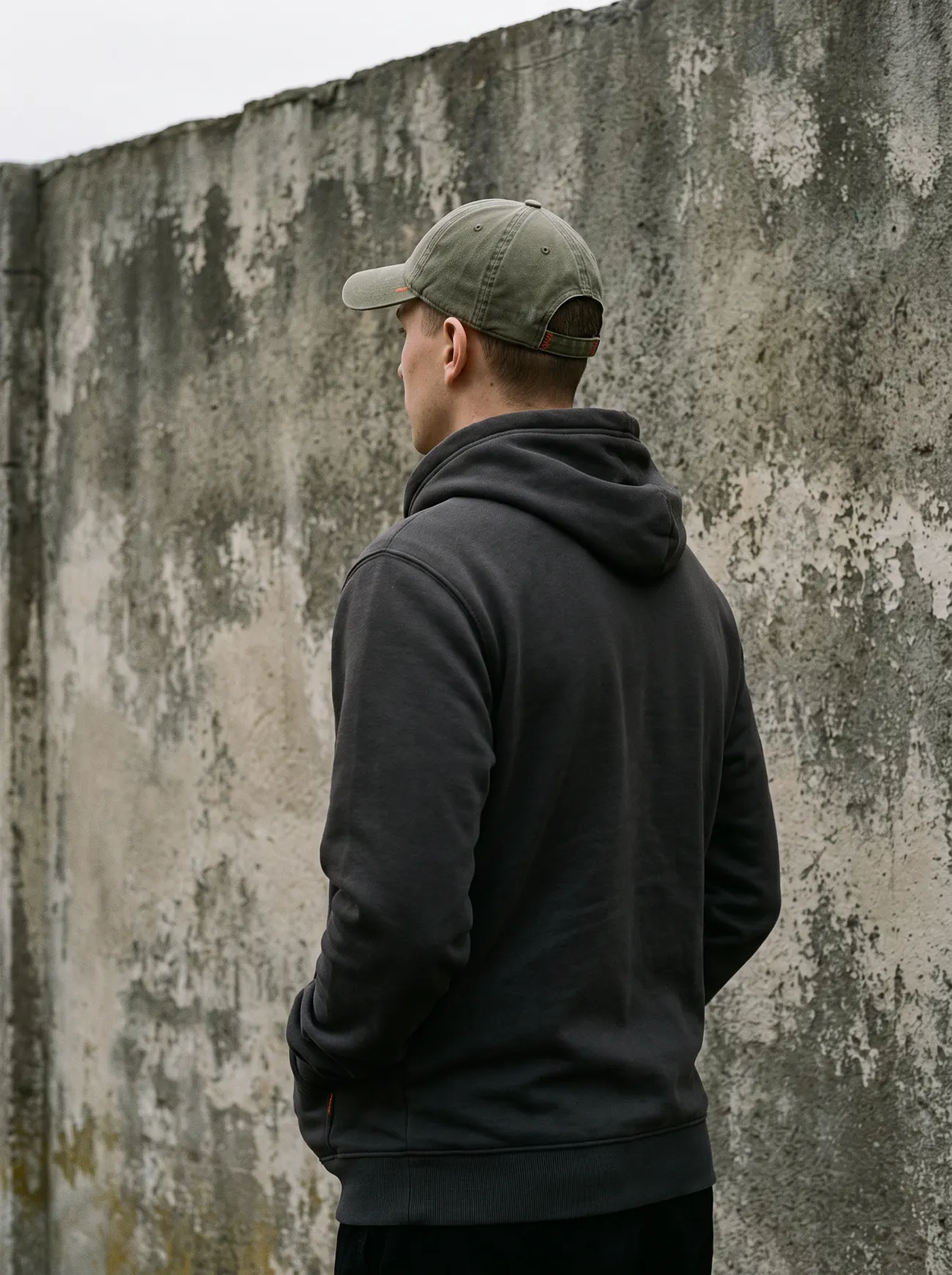 A person seen from behind in a charcoal hoodie and an olive cap against a concrete wall