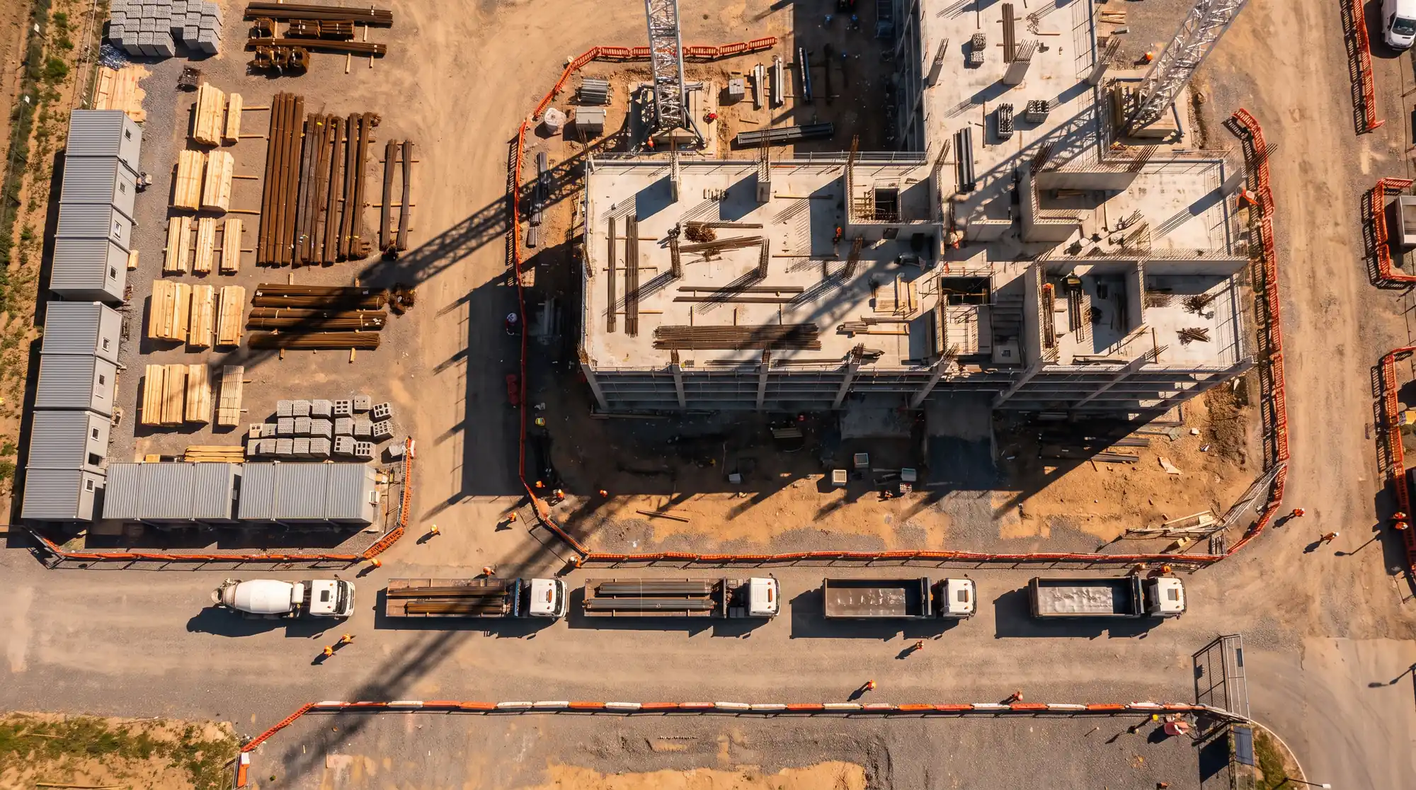 A drone view straight down on a building site with two tower cranes, a laydown yard and a line of delivery trucks queued to the gate