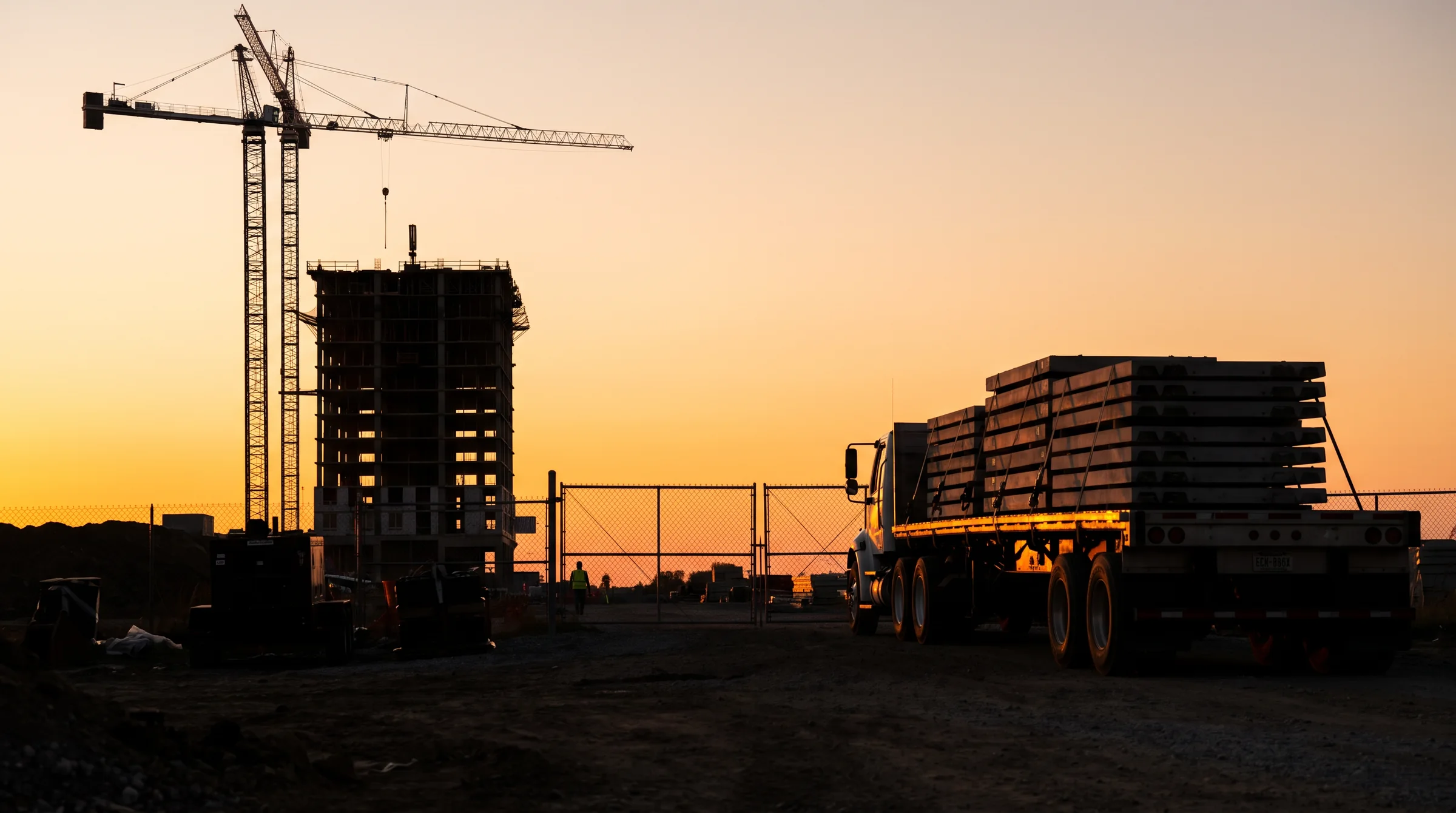 A tower crane, a half-built concrete frame and a flatbed truck loaded with precast panels at a site gate, all black silhouettes against an orange sunset