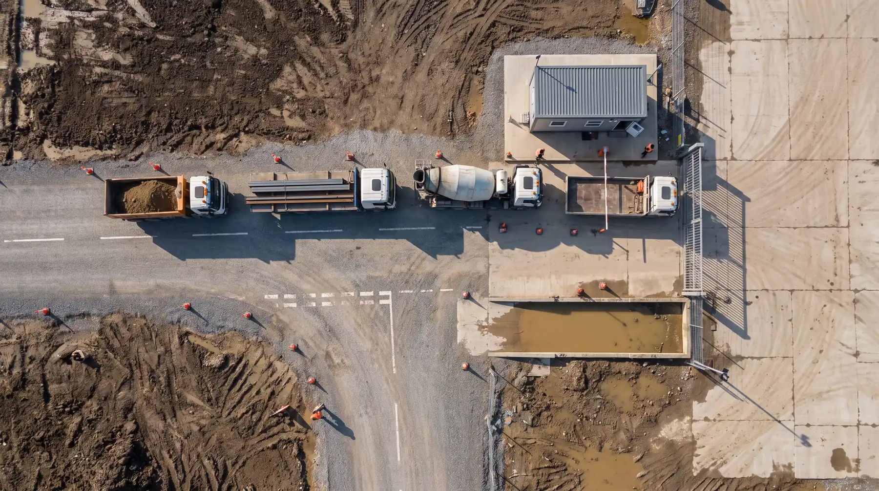 A drone view straight down on a site entrance with a boom barrier, a gatehouse and delivery trucks queued on a gravel road