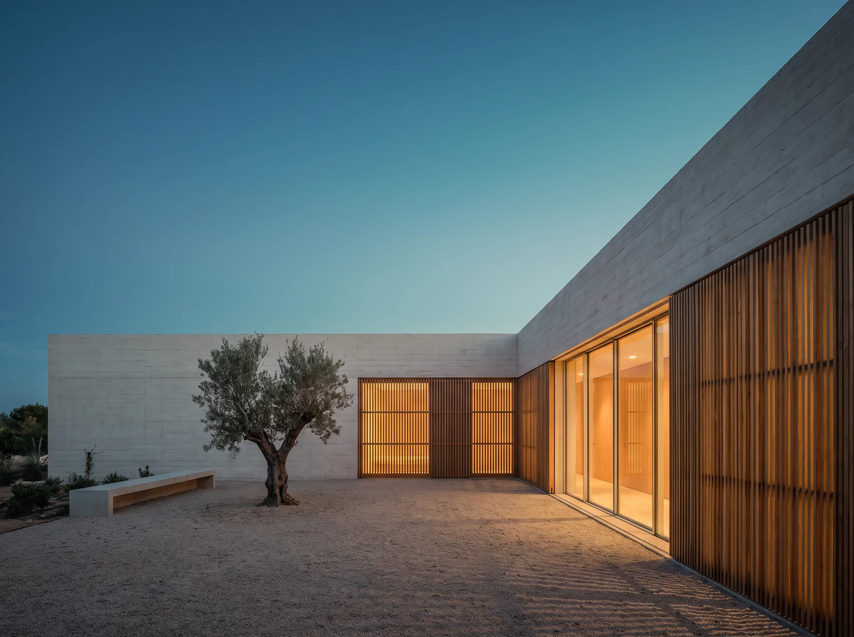 A low courtyard house of pale concrete and oak slats at blue hour with warm light in the windows