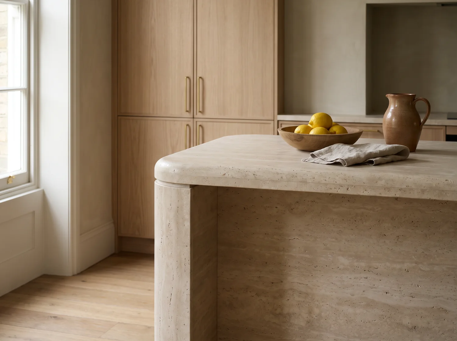 A kitchen island in honed travertine with ash cabinets, brass handles and a bowl of lemons