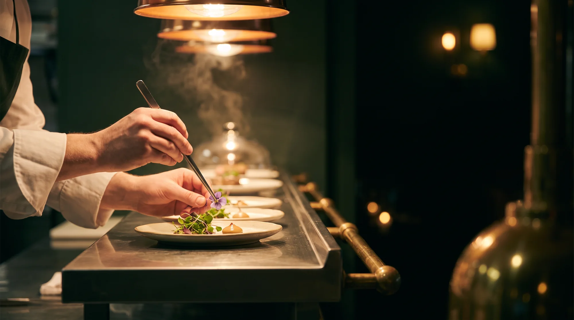 A chef's hands in a white cuff placing a flower with tweezers onto a plate at a steel pass under warm lamps