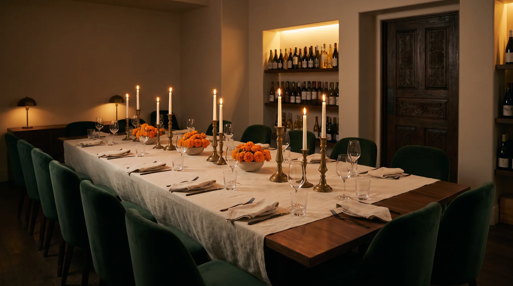 A long walnut table set with brass candlesticks and marigolds in a private dining room with a lit wall of wine bottles