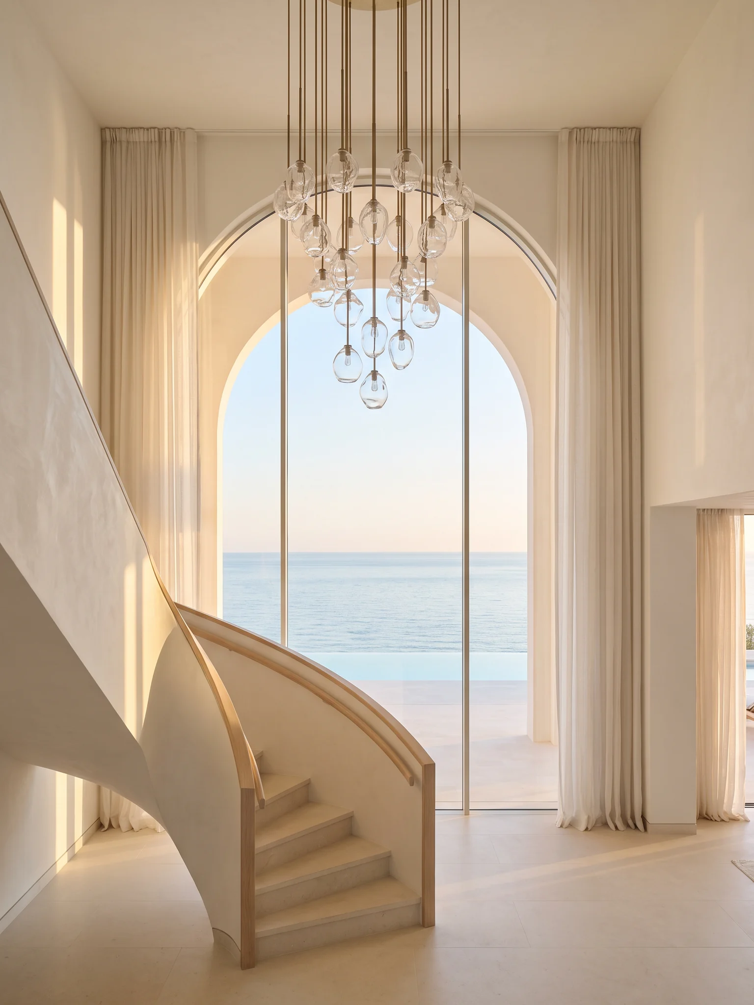 A double-height entrance hall with a tall arched window, a glass and bronze chandelier and a curved limestone staircase