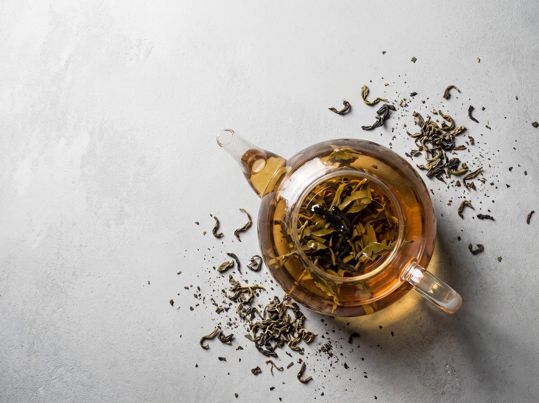 A glass teapot of golden tea with loose leaves scattered on a pale surface, seen from above