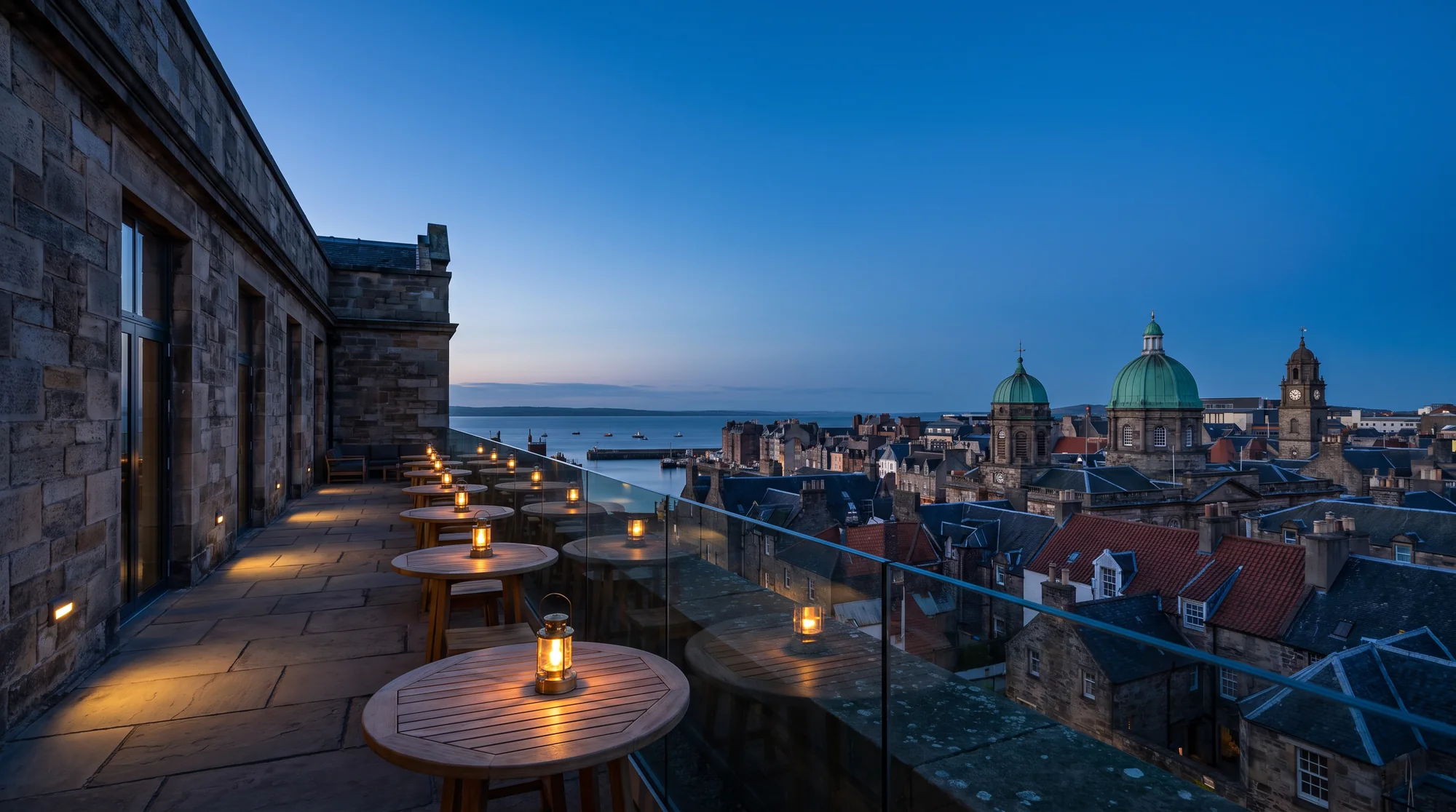 The rooftop terrace at blue hour above a harbour city of tiled roofs, a green dome and a clock tower