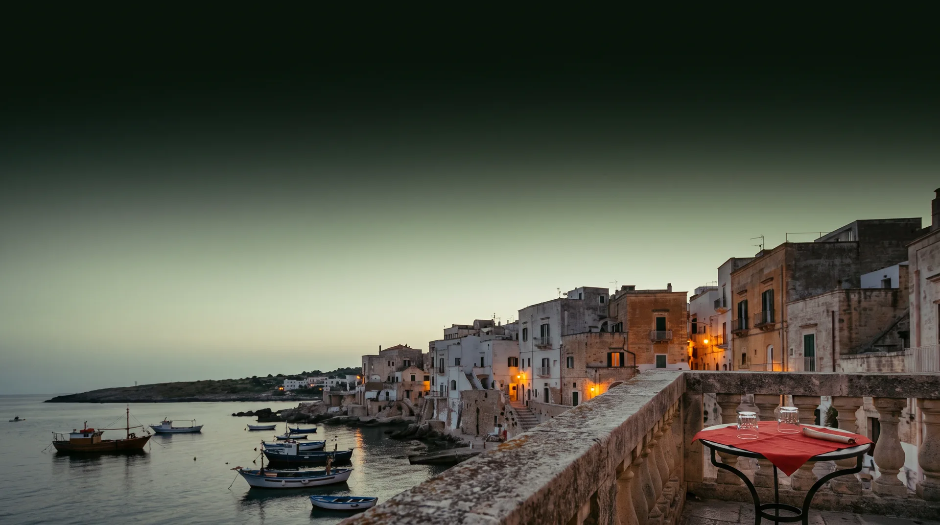 A hotel terrace at dusk above a quiet harbour town, with a small table set for two