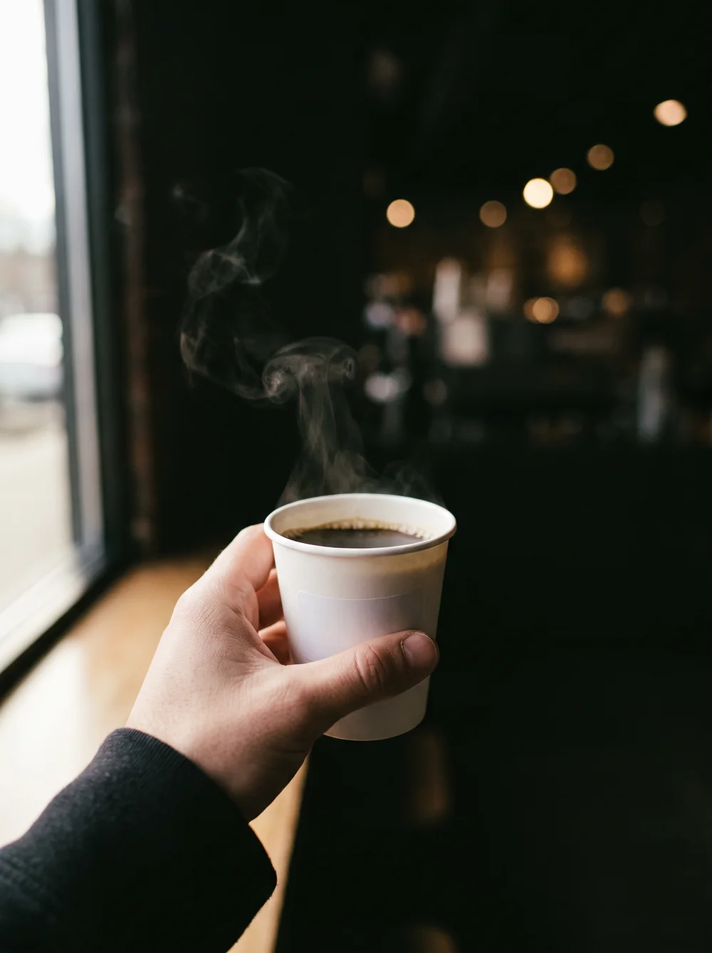 A hand holding a small white paper cup of black coffee in a dark cafe