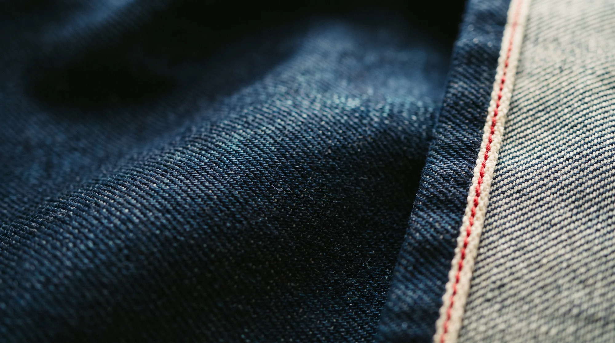 Dark indigo denim in low light, with a pale cotton selvedge edge and a red thread line