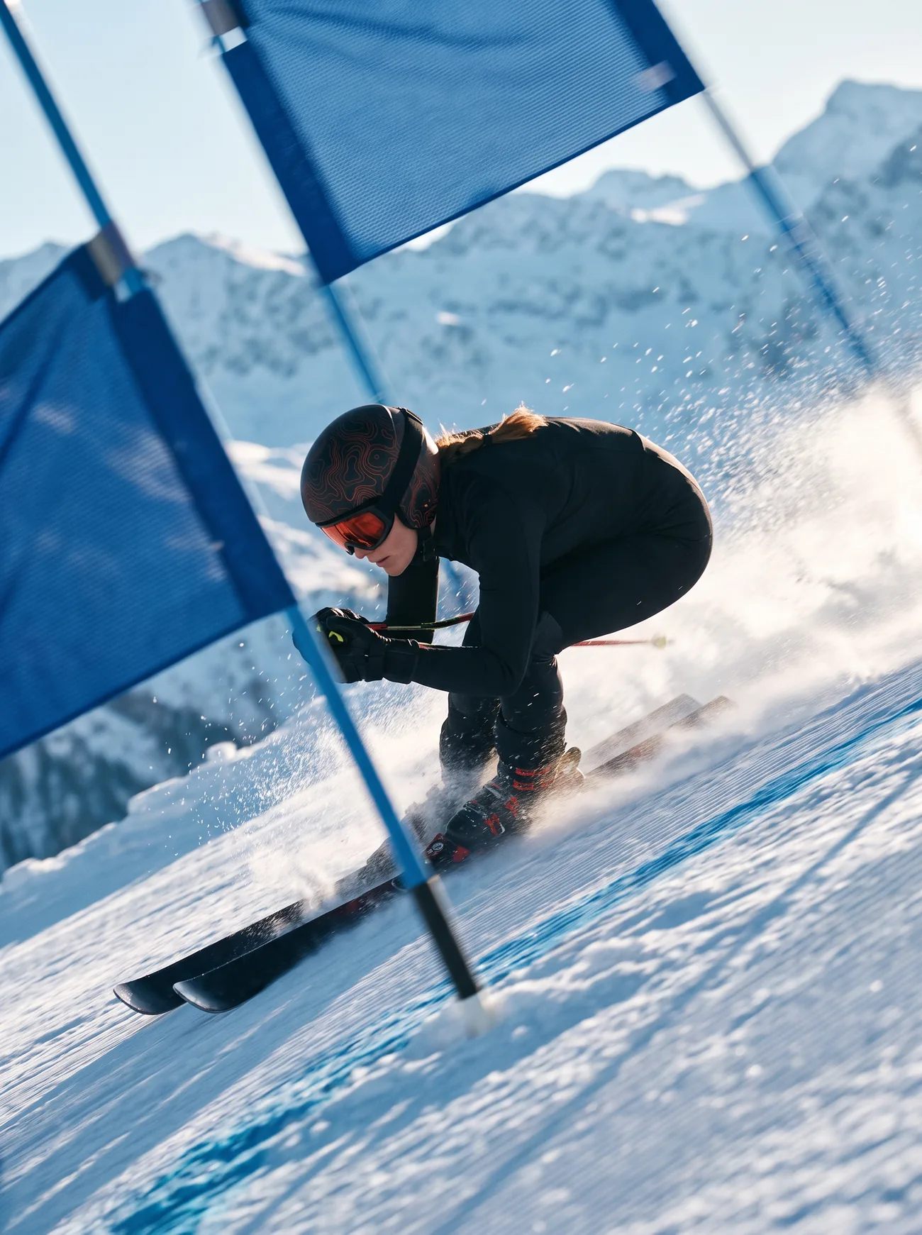 Kaia in a low tuck at speed past a blue gate, snow spraying off her skis