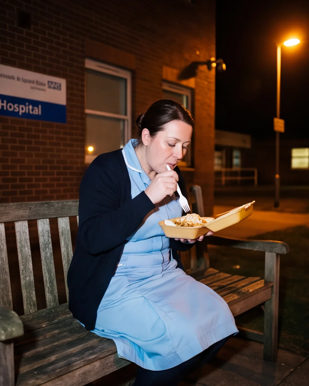 Night Shift, photographs by Corin Vasko: A nurse in a pale uniform eating from a foil tray on a bench at night