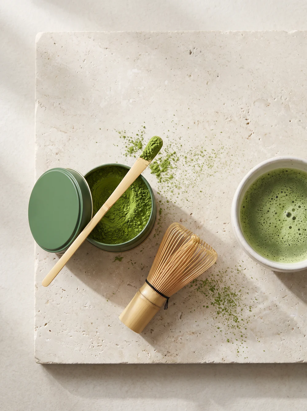A dark green tin of bright matcha powder with a bamboo scoop and whisk on a pale stone slab
