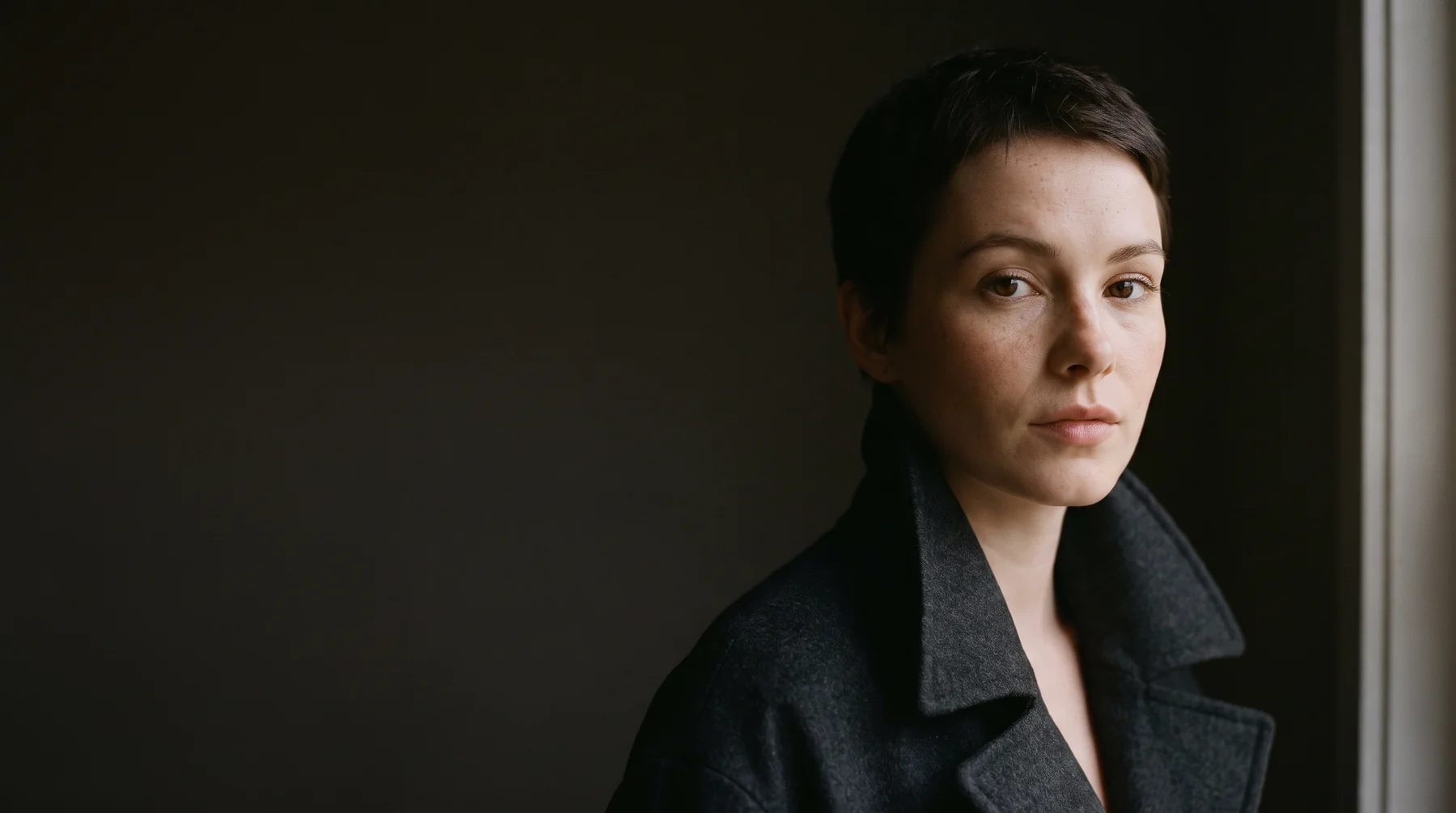A close portrait of a woman in a charcoal coat, lit from a window on one side