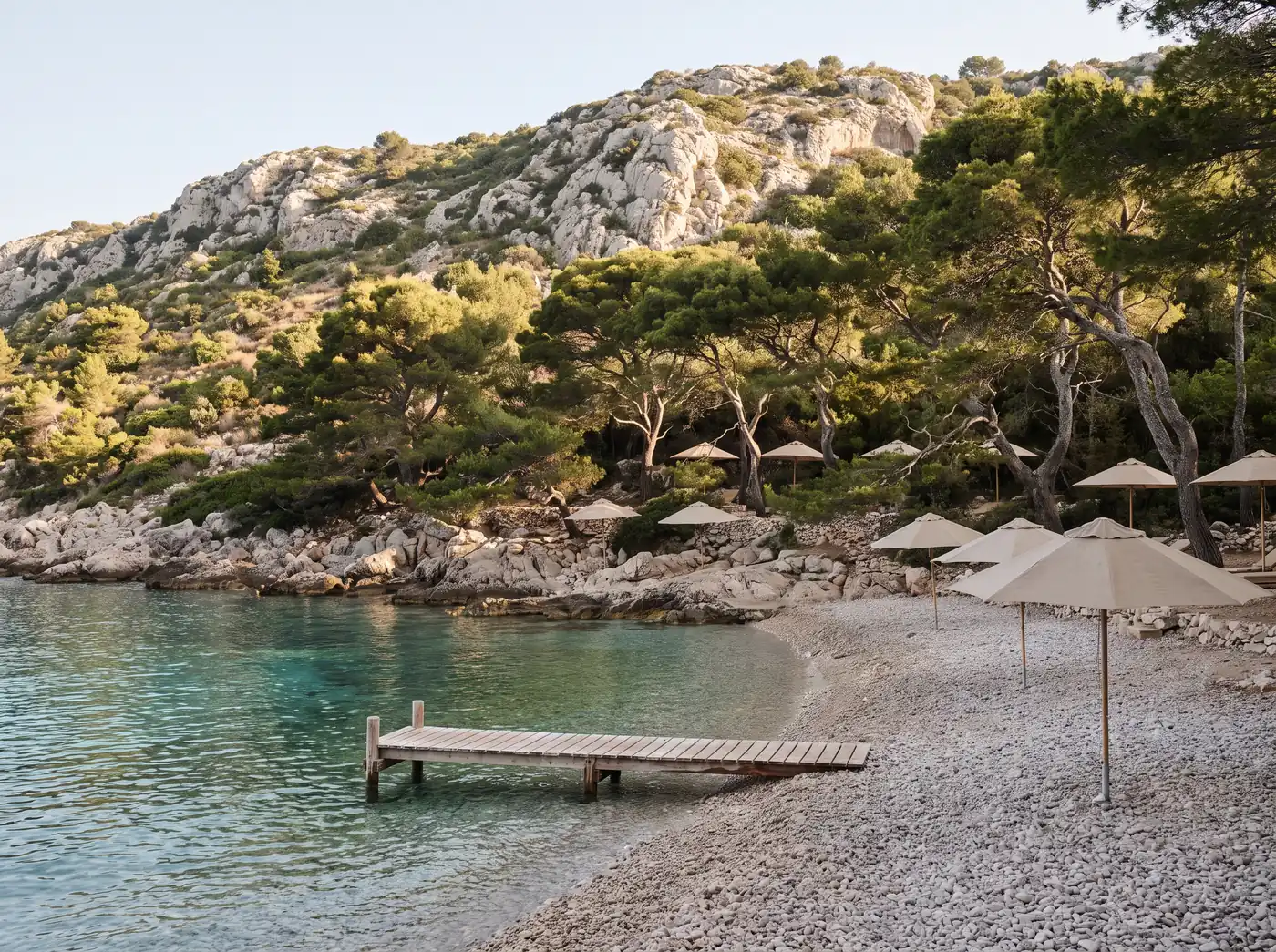 A quiet pebble cove with clear turquoise water, umbrella pines and a wooden jetty