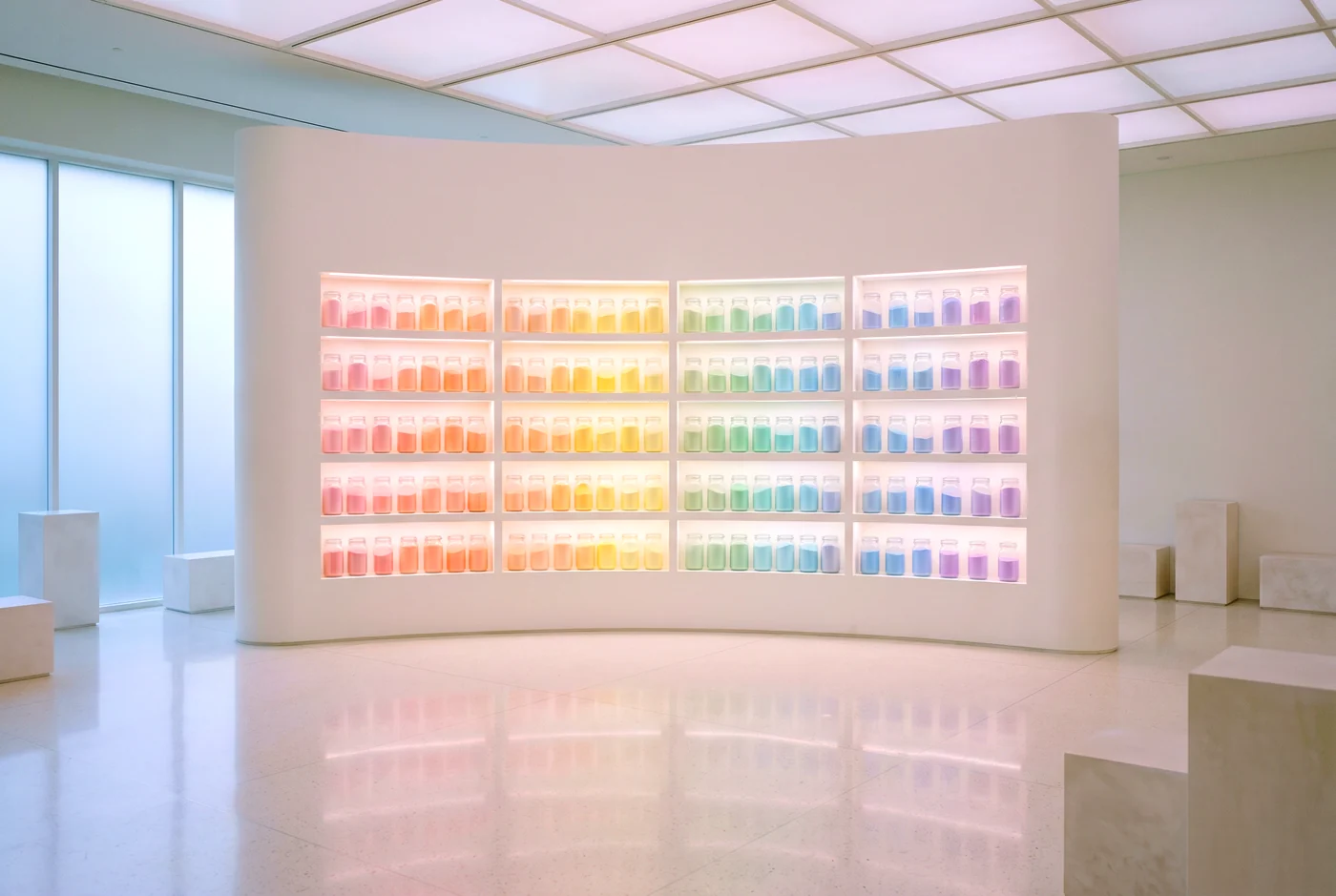 A long white wall of shelves holding hundreds of glass jars of pastel pigment, one section lit from within